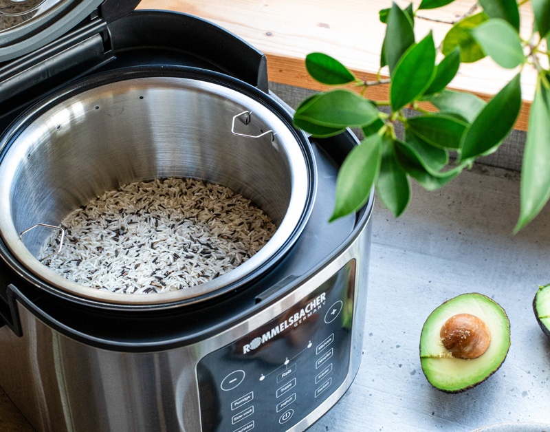 ROMMELSBACHER Multi-Reiskocher MRK 650 mit geöffnetem Deckel und frisch gekochtem Reis. Daneben eine aufgeschnittene Avocado und grüne Dekozweige.