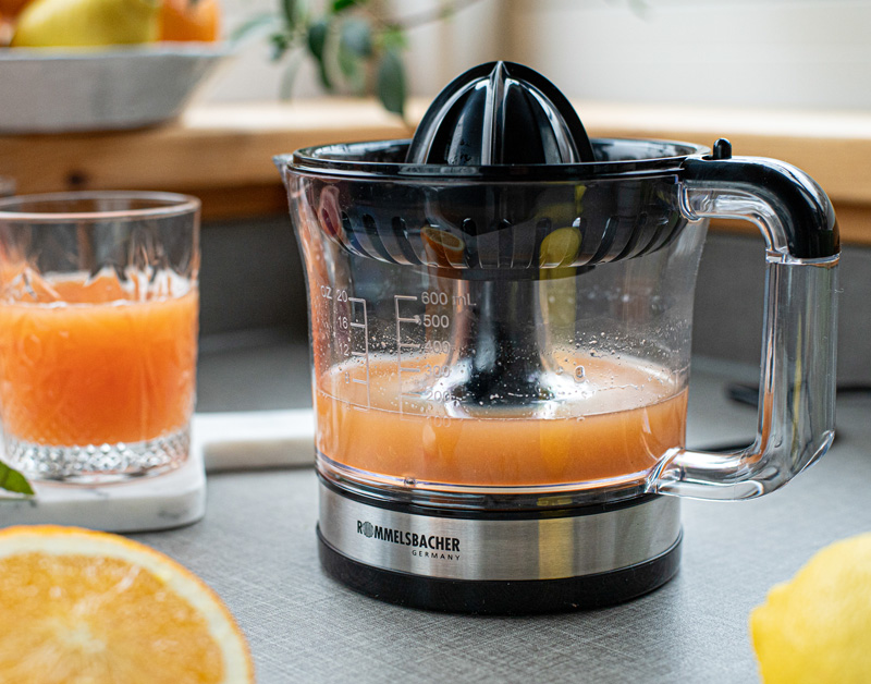 ROMMELSBACHER Zitruspresse ZP 50 beim Entsaften einer Orange. Daneben ein Glas mit frischem Saft und angeschnittene Zitrusfrüchte auf dem Tisch.
