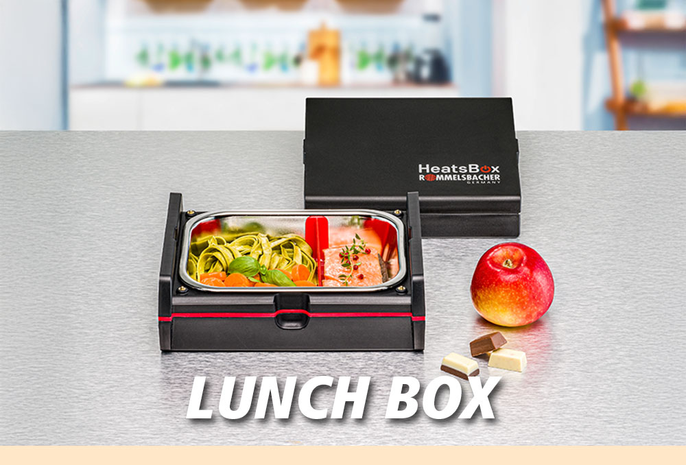 Eine elektrische Lunchbox mit geöffnetem Deckel, gefüllt mit Gemüse und Fleisch, daneben ein Apfel und Schokoladenwürfel. Im Bild steht in weißer Schrift: „LUNCH BOX“.