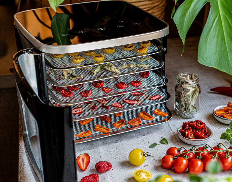 ROMMELSBACHER Dörrautomat DA 450 mit fünf Einschüben, belegt mit getrocknetem Dörrgut. Daneben frisches Gemüse und ein Glas mit getrocknetem Dörrgut.